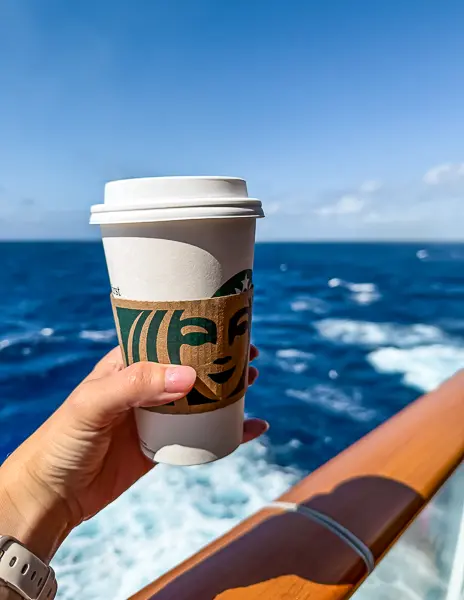 A hand holding a Starbucks coffee cup against a backdrop of the ocean view from the Norwegian Getaway.