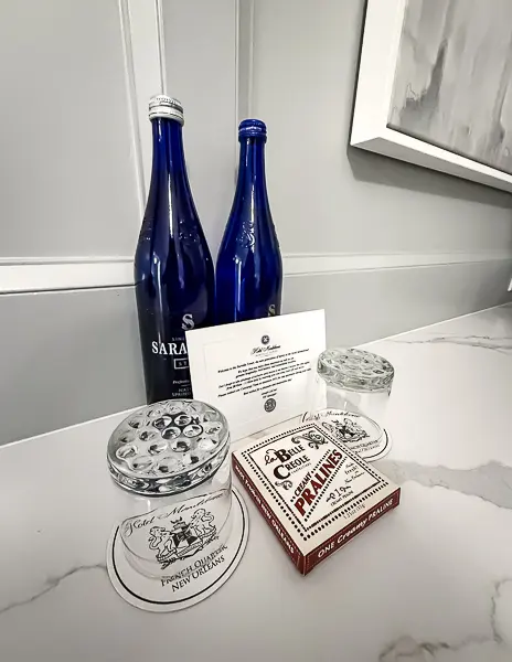 A welcoming display in the King Room at Hotel Monteleone, featuring two bottles of Saratoga spring water, coasters with the hotel's logo, a box of pralines, and a personalized note, showcasing thoughtful hospitality.