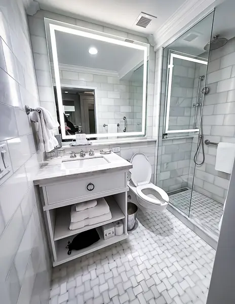 A modern and elegant bathroom in the Iberville King Room at Hotel Monteleone, featuring marble tile, a sleek vanity with open shelving, a backlit mirror, and a glass-enclosed rainfall shower for a luxurious experience.