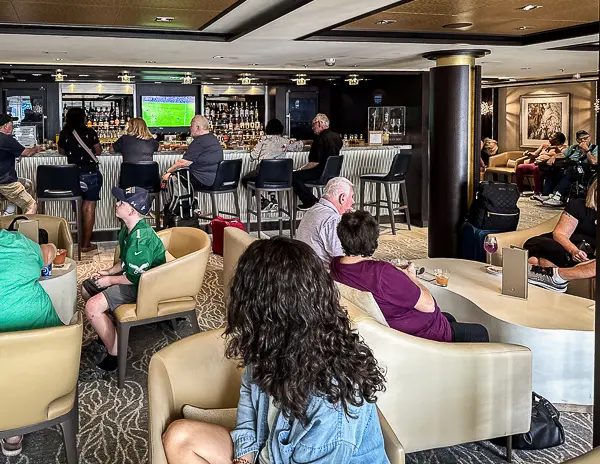 Guests relaxing in The Haven lounge on the Norwegian Getaway, with a bar area and bartenders serving drinks in the background.