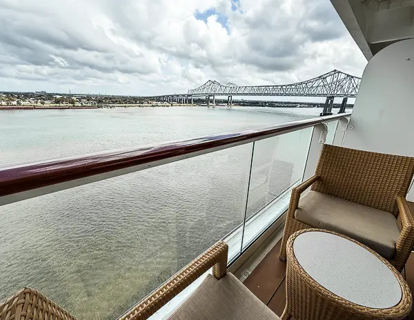 A view from the balcony of the Norwegian Getaway Haven Penthouse Suite 14138, featuring wicker chairs, a small table, and a scenic view of the Mississippi River and a bridge in New Orleans