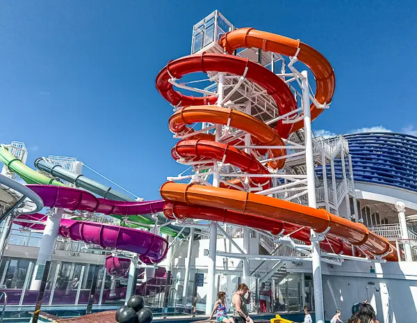 Colorful water slides on the Norwegian Getaway, featuring twists and turns in orange, red, and purple, with cruise guests enjoying the pool deck below on a sunny day.