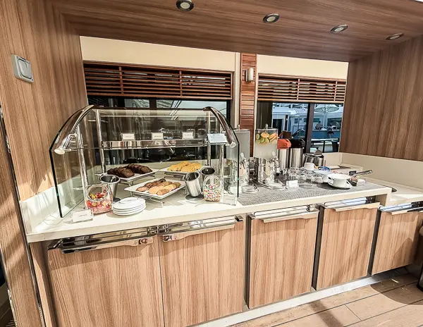 Snack and drink station near The Haven pool on the Norwegian Getaway, featuring pastries, fresh fruit, and a selection of beverages in a stylish wooden counter setup.