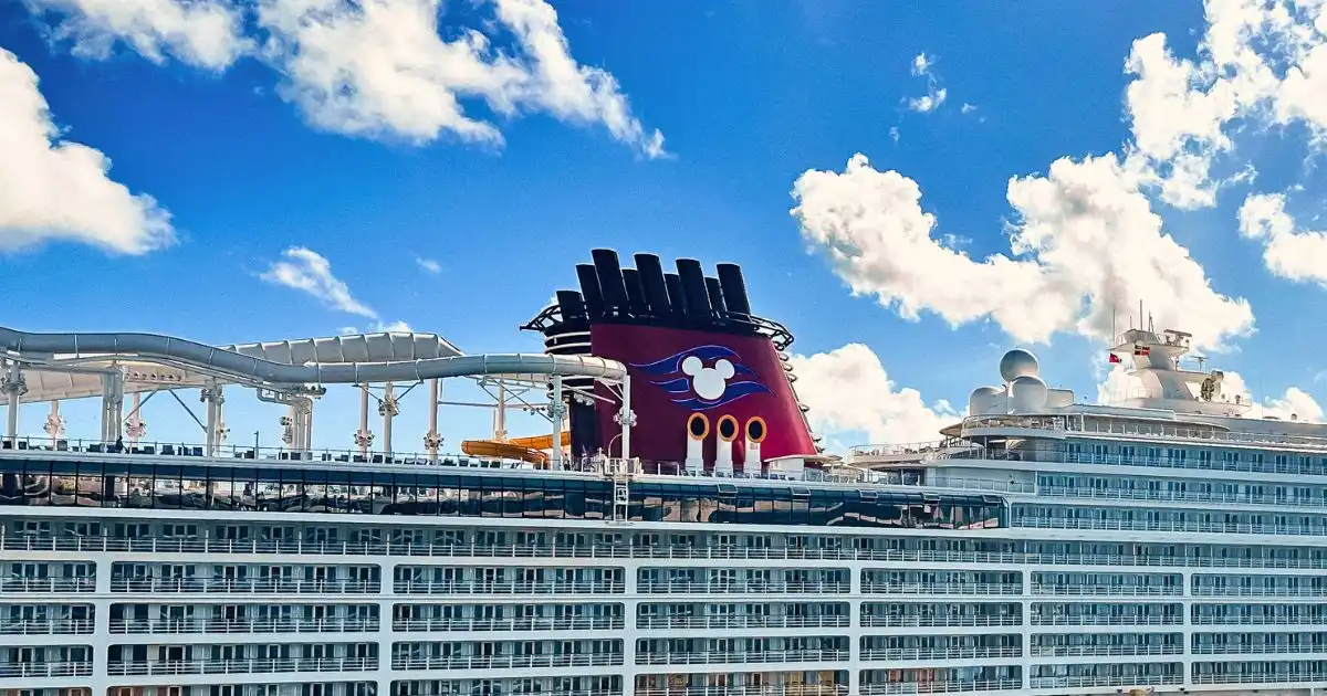 A Disney Cruise Line ship featuring its signature red funnel with Mickey Mouse logo, along with an onboard waterslide, set against a bright blue sky with scattered clouds.