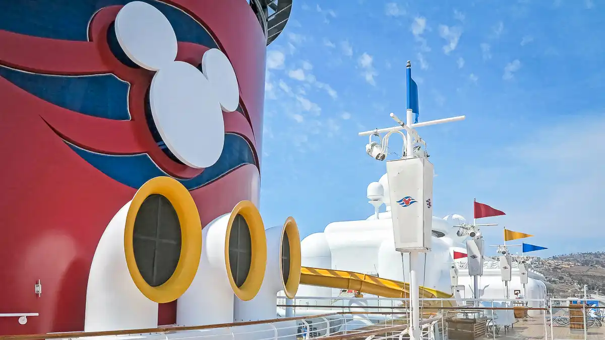 Disney Cruise Line ship deck featuring the iconic red funnel with Mickey Mouse logo, yellow portholes, and colorful flags against a bright blue sky.