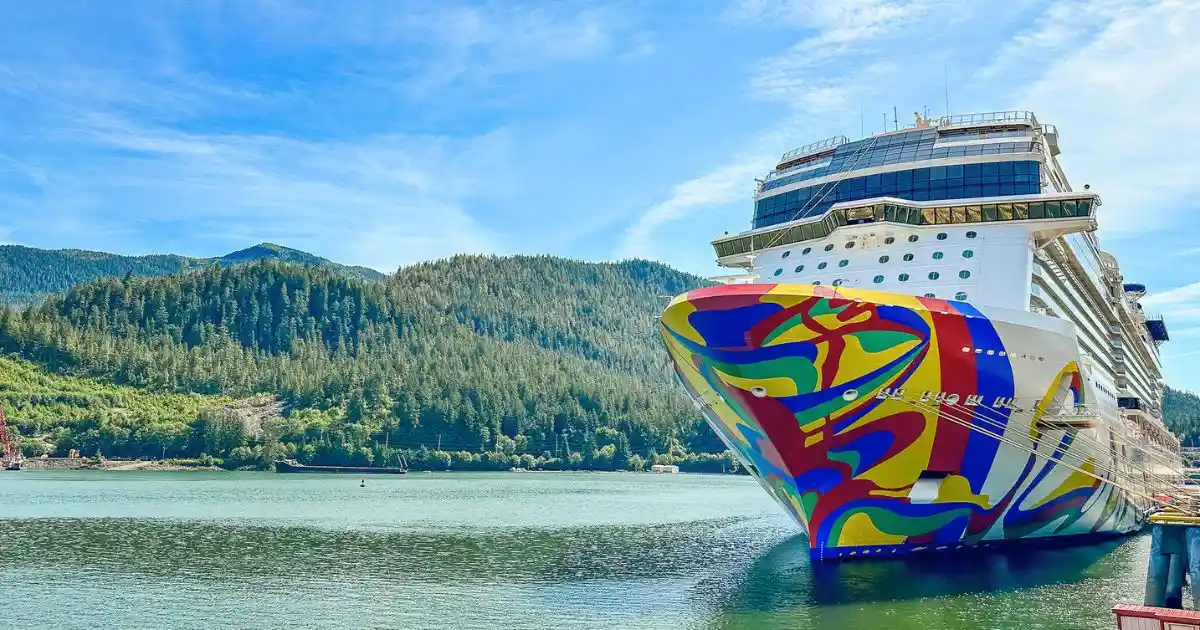 The Norwegian Encore cruise ship, adorned with vibrant, abstract hull art, sails under a clear blue sky. The vessel features multiple decks with rows of balconies, a prominent observation lounge at the bow, and the distinctive Encore Speedway go-kart track visible on the top deck.