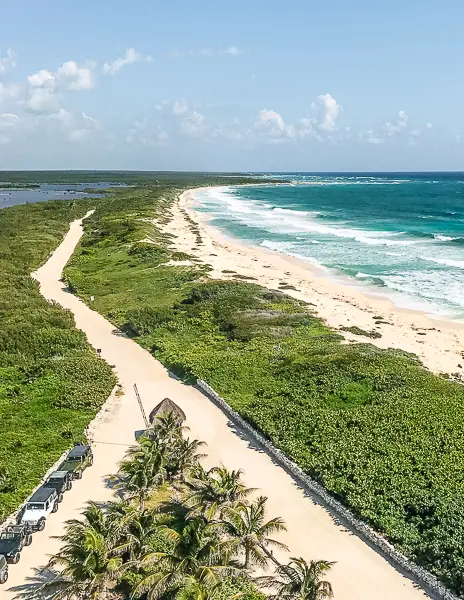 Scenic aerial view of Punta Sur Eco Park in Cozumel, featuring a sandy coastal road, lush greenery, and a stretch of untouched beach with turquoise waves.