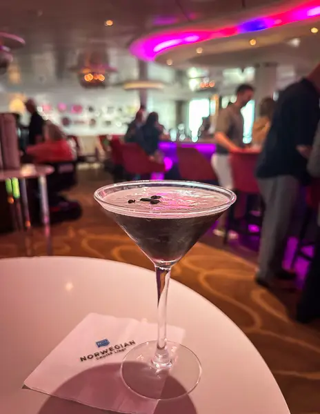 A freshly made espresso martini served in a classic martini glass on a Norwegian Cruise Line ship, placed on a branded napkin with a lively cruise bar in the background.