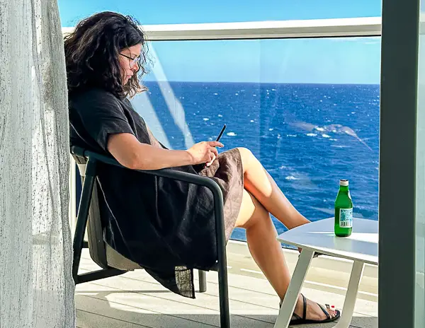 A woman (kathy) relaxes on a private cruise ship balcony, enjoying ocean views while using a tablet, with a bottle of sparkling water on the table beside her.