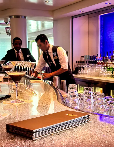 Bartenders preparing drinks at a stylish martini bar on a Norwegian Cruise Line ship, featuring a sleek counter, cocktail glasses, and an illuminated bar backdrop.