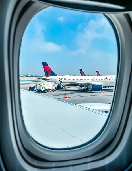 View from an airplane window showing Delta Air Lines planes parked at the gate, with ground crew preparing for takeoff under a bright blue sky.