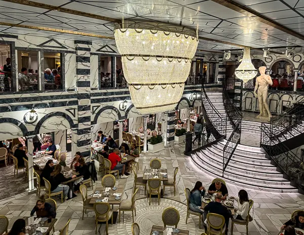 Michelangelo Restaurant on Carnival Firenze, featuring a grand staircase, chandeliers, marble-style decor, and guests dining across two levels.