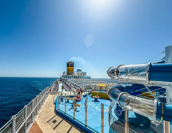 View of Carnival Firenze’s upper deck with waterslides and kids’ splash zone in the foreground, and the ship’s Costa-style funnel in the distance under a clear blue sky.