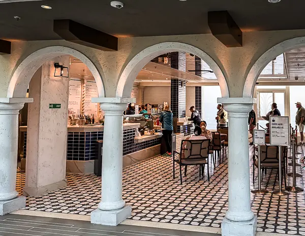 JavaBlue Café on Carnival Firenze viewed through arched entryways, with guests ordering coffee and sitting at tables on a patterned tile floor.